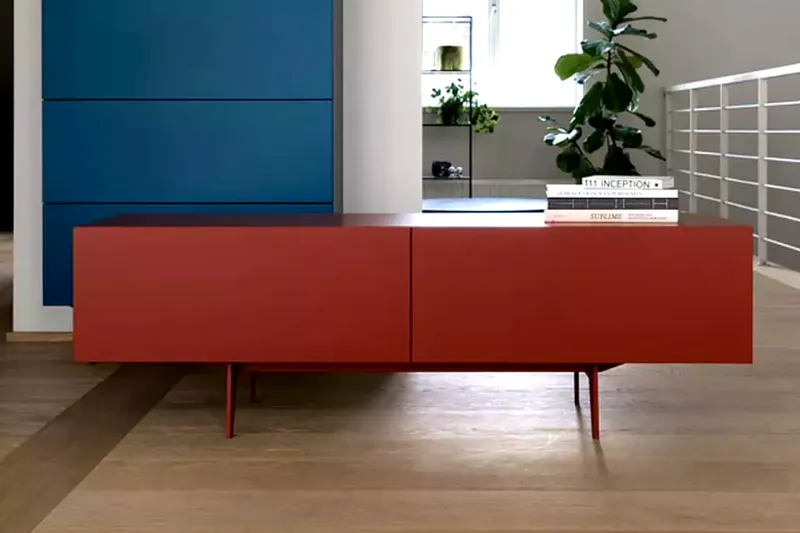 Rotes Sideboard mit zwei Schubladen und Metallbeinen vor blauem Wandpaneel und Regalen mit Pflanzen und Büchern.