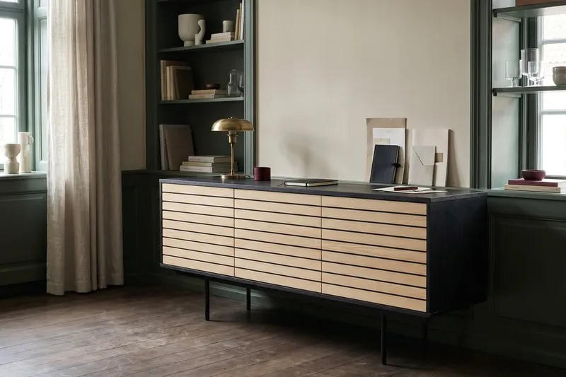 Sideboard aus hellem Eichenfurnier mit schwarzem Rahmen und Schubladen, dahinter schwarze Wandregale mit Büchern und Deko.