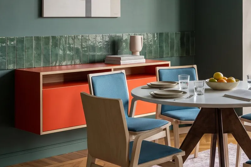 Rotes Sideboard mit offenen Fächern und orangefarbenem Rückwand neben rundem Esstisch mit blauen Stühlen in Wohnraum.