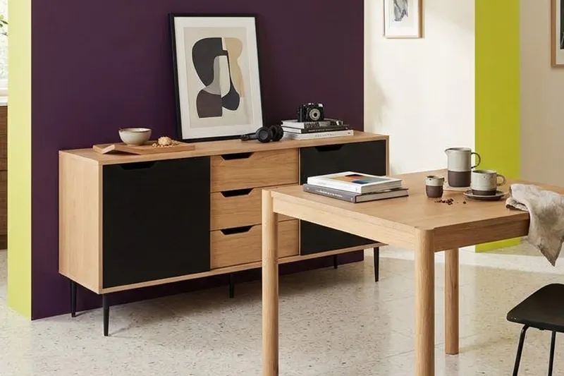 Sideboard aus Eiche mit schwarzen Schubkästen und Türen auf Metallbeinen, daneben Esstisch mit Geschirr und Büchern, abstrakt